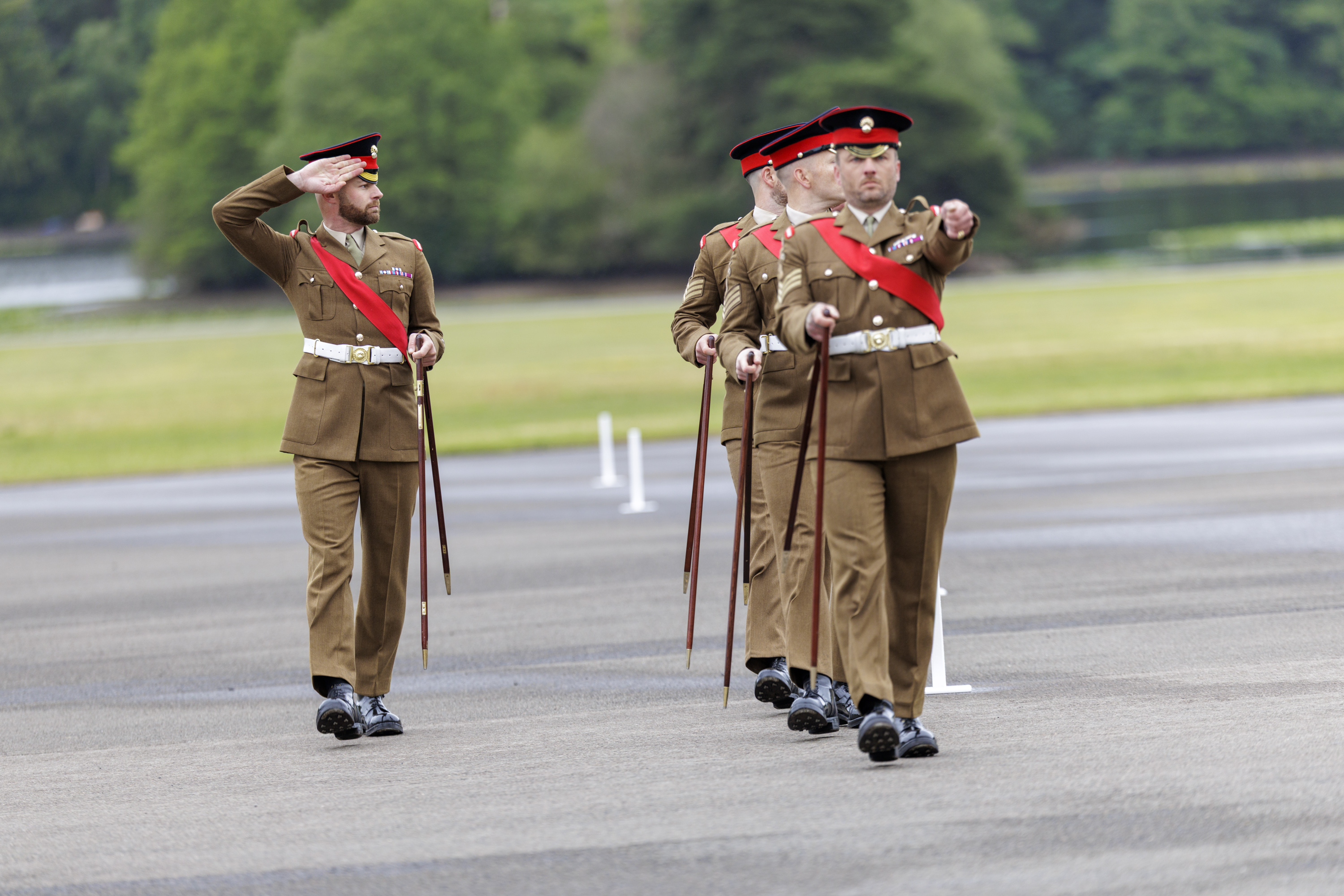 A group of uniformed soldiers is marching in formation across a paved area. They are wearing matching brown uniforms with red sashes and are holding ceremonial swords. One of the soldiers at the front is saluting. The background features greenery and blurred trees.