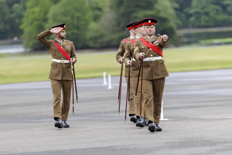 A group of uniformed soldiers is marching in formation across a paved area. They are wearing matching brown uniforms with red sashes and are holding ceremonial swords. One of the soldiers at the front is saluting. The background features greenery and blurred trees.