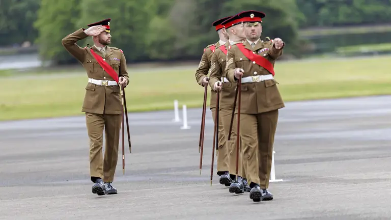 A group of uniformed soldiers is marching in formation across a paved area. They are wearing matching brown uniforms with red sashes and are holding ceremonial swords. One of the soldiers at the front is saluting. The background features greenery and blurred trees.