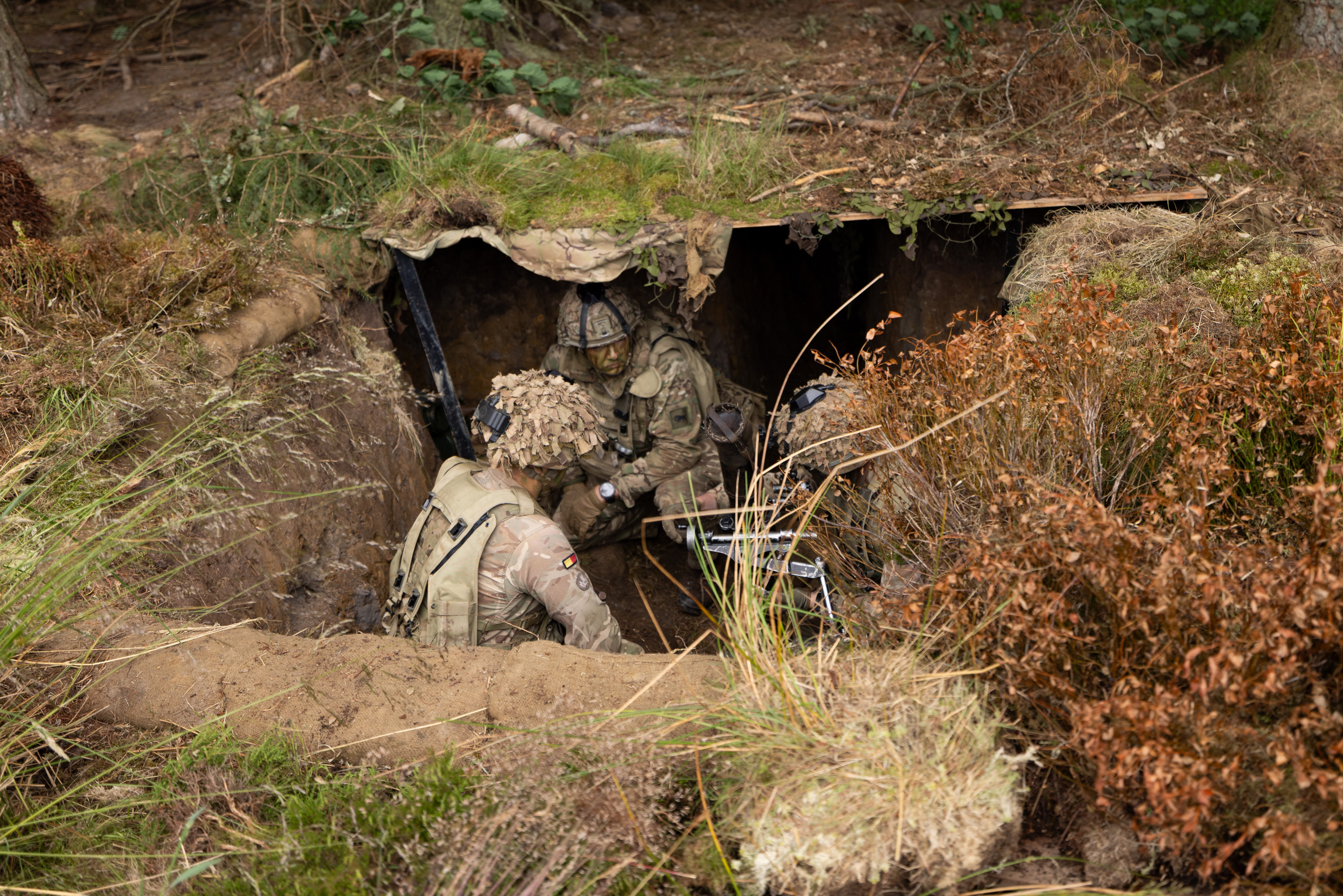 Military personnel in camouflage gear are positioned in a small, camouflaged bunker, surrounded by foliage.