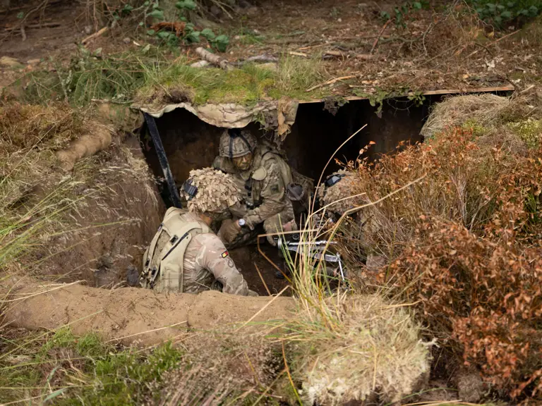 Military personnel in camouflage gear are positioned in a small, camouflaged bunker, surrounded by foliage.