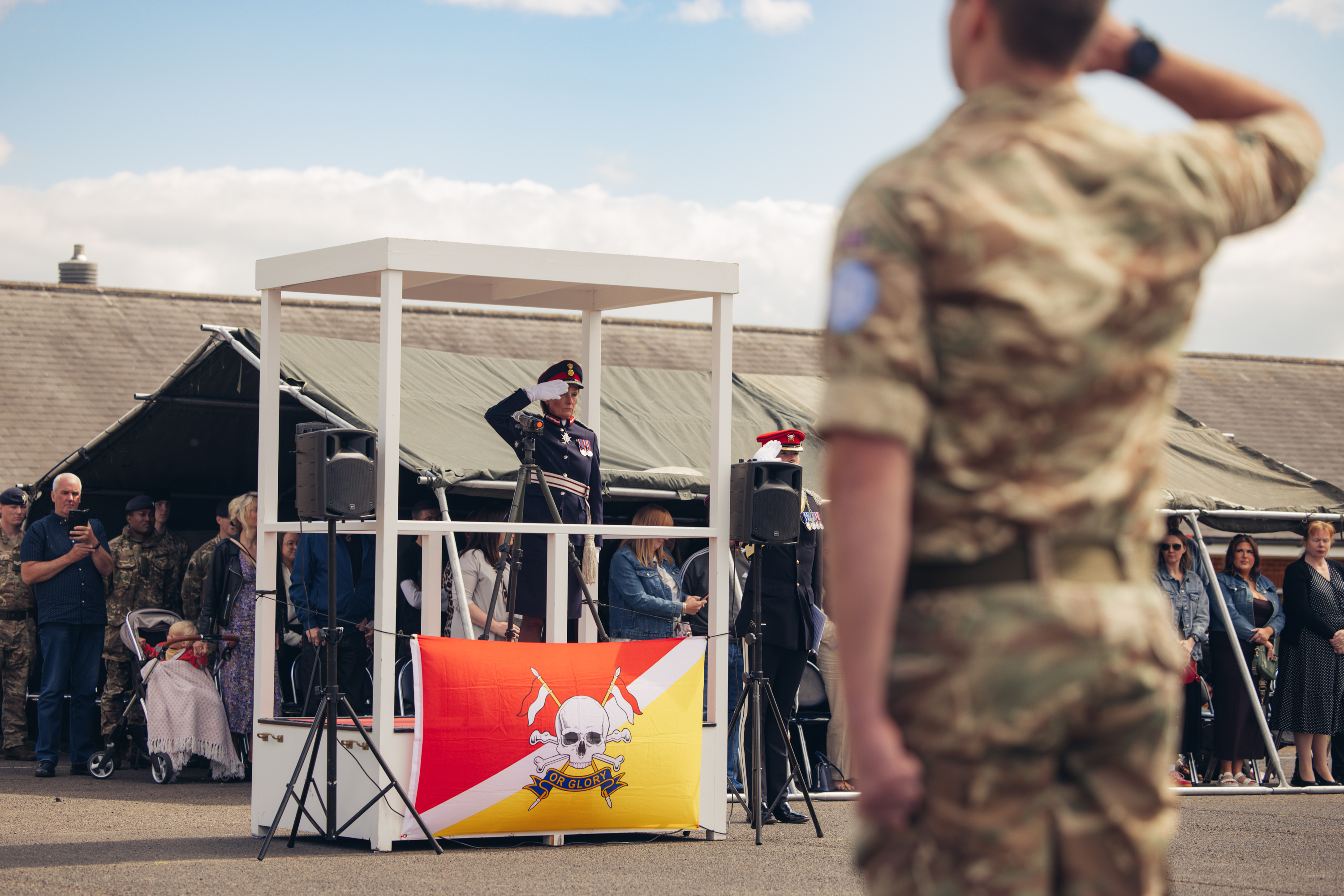 A woman in ceremonial attire salutes from a stage as an audience looks on from behind the stage. 