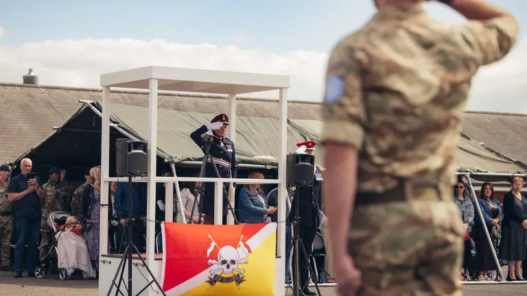 A woman in ceremonial attire salutes from a stage as an audience looks on from behind the stage.