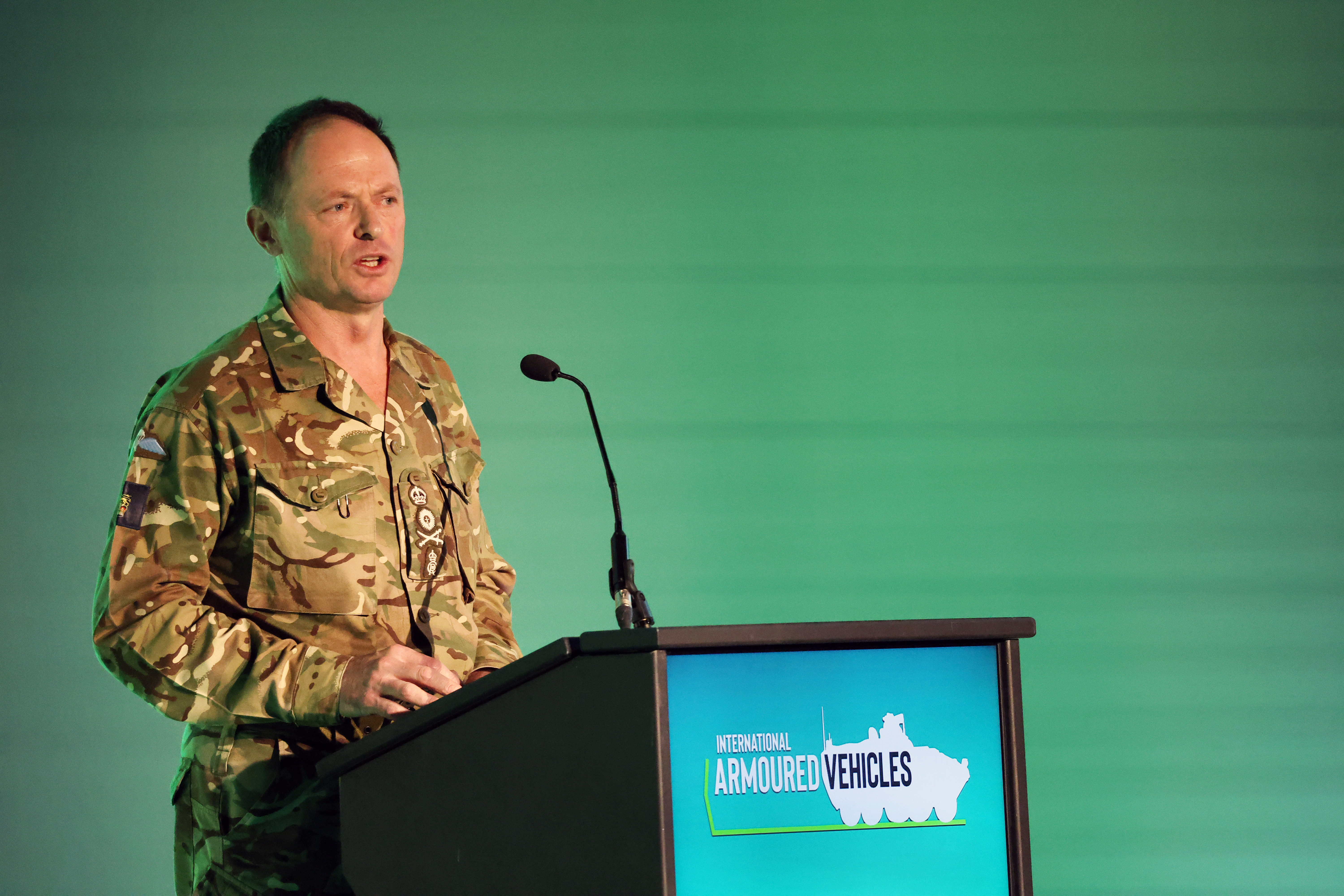 Military personnel in camouflage uniform speaking at a podium with an International Armoured Vehicles event sign.
