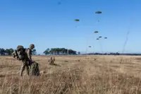 Soldiers in camouflage gear prepare equipment in a dry field while multiple parachutes descend from the clear blue sky.
