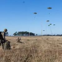 Soldiers in camouflage gear prepare equipment in a dry field while multiple parachutes descend from the clear blue sky.