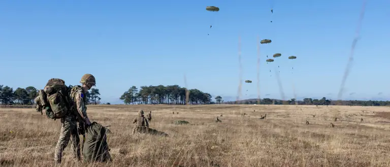 Soldiers in camouflage gear prepare equipment in a dry field while multiple parachutes descend from the clear blue sky.