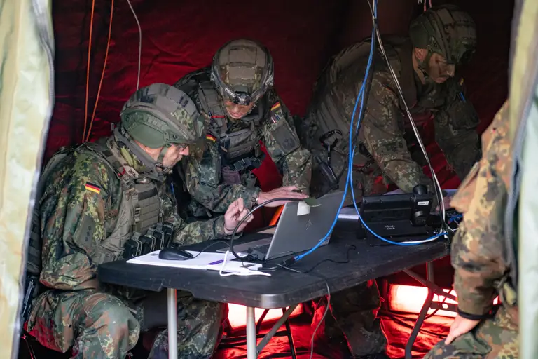 Two German soldiers lean over a laptop within a green tent.