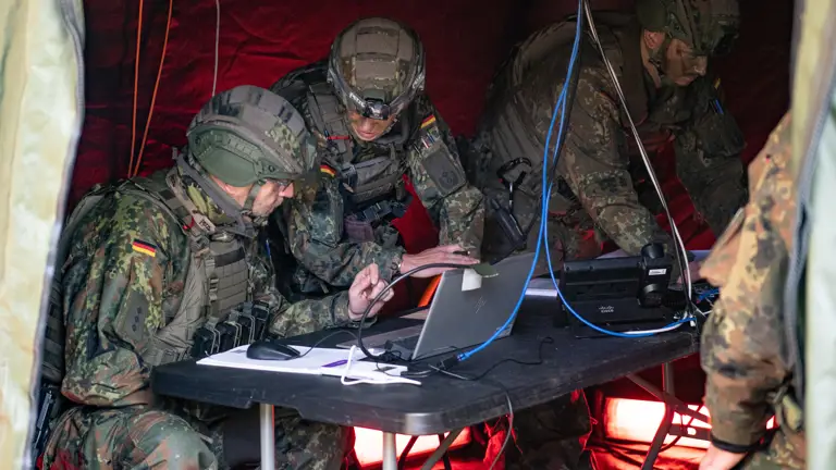Two German soldiers lean over a laptop within a green tent.