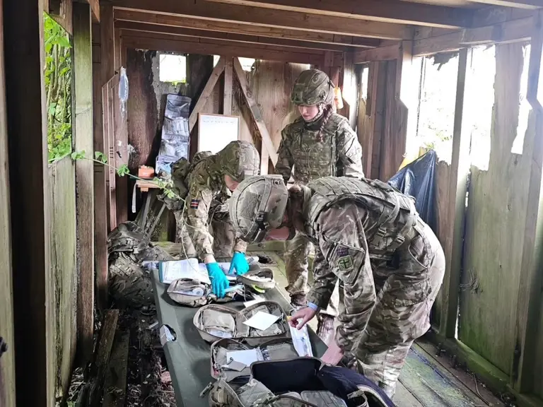 Soldiers in camouflage uniforms examine medical kits on a table in a wooden shelter. The scene conveys focus and teamwork, with a rural backdrop.