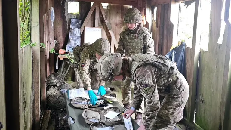 Soldiers in camouflage uniforms examine medical kits on a table in a wooden shelter. The scene conveys focus and teamwork, with a rural backdrop.