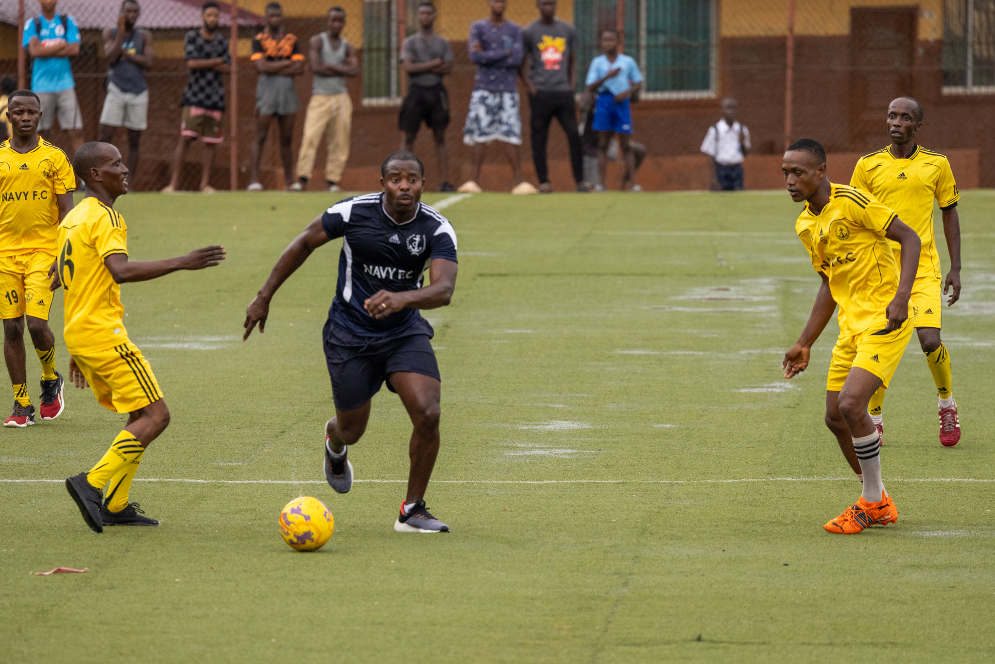 A man wearing a black football kit is pictured dribbling a football past a team wearing yellow kit. 
