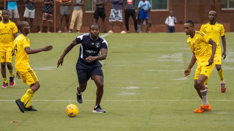 A man wearing a black football kit is pictured dribbling a football past a team wearing yellow kit.