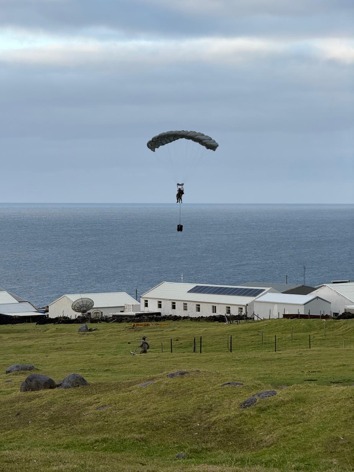 Paratrooper descends over grassy field with ocean backdrop, landing near small white buildings. Overcast sky creates a calm, muted atmosphere.