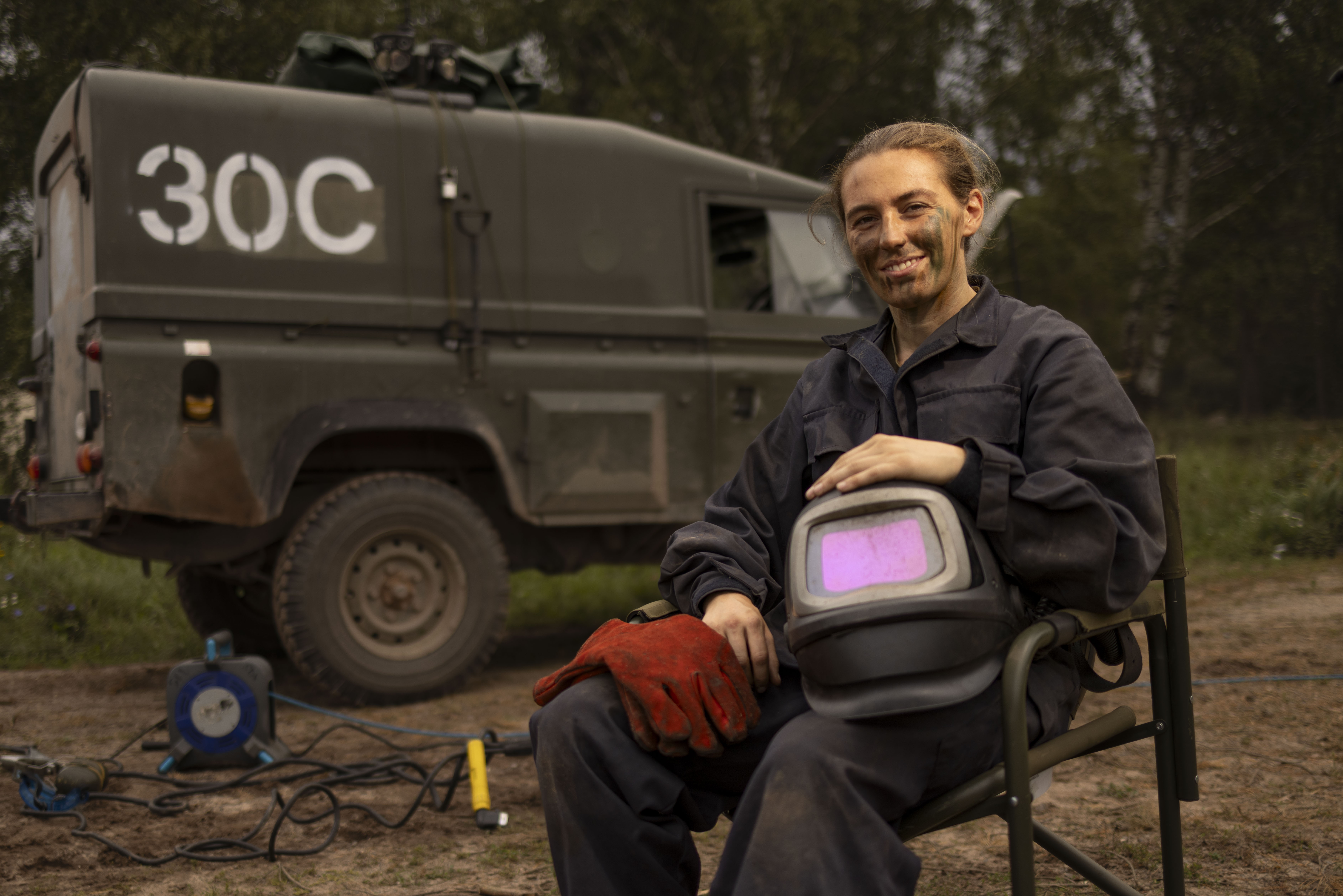 Person in dark workwear holding a welding helmet and red gloves, seated outdoors near a military-style vehicle.