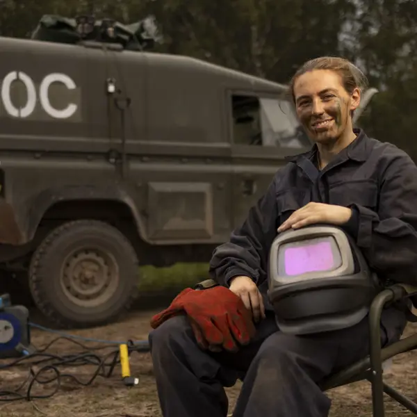 Person in dark workwear holding a welding helmet and red gloves, seated outdoors near a military-style vehicle.