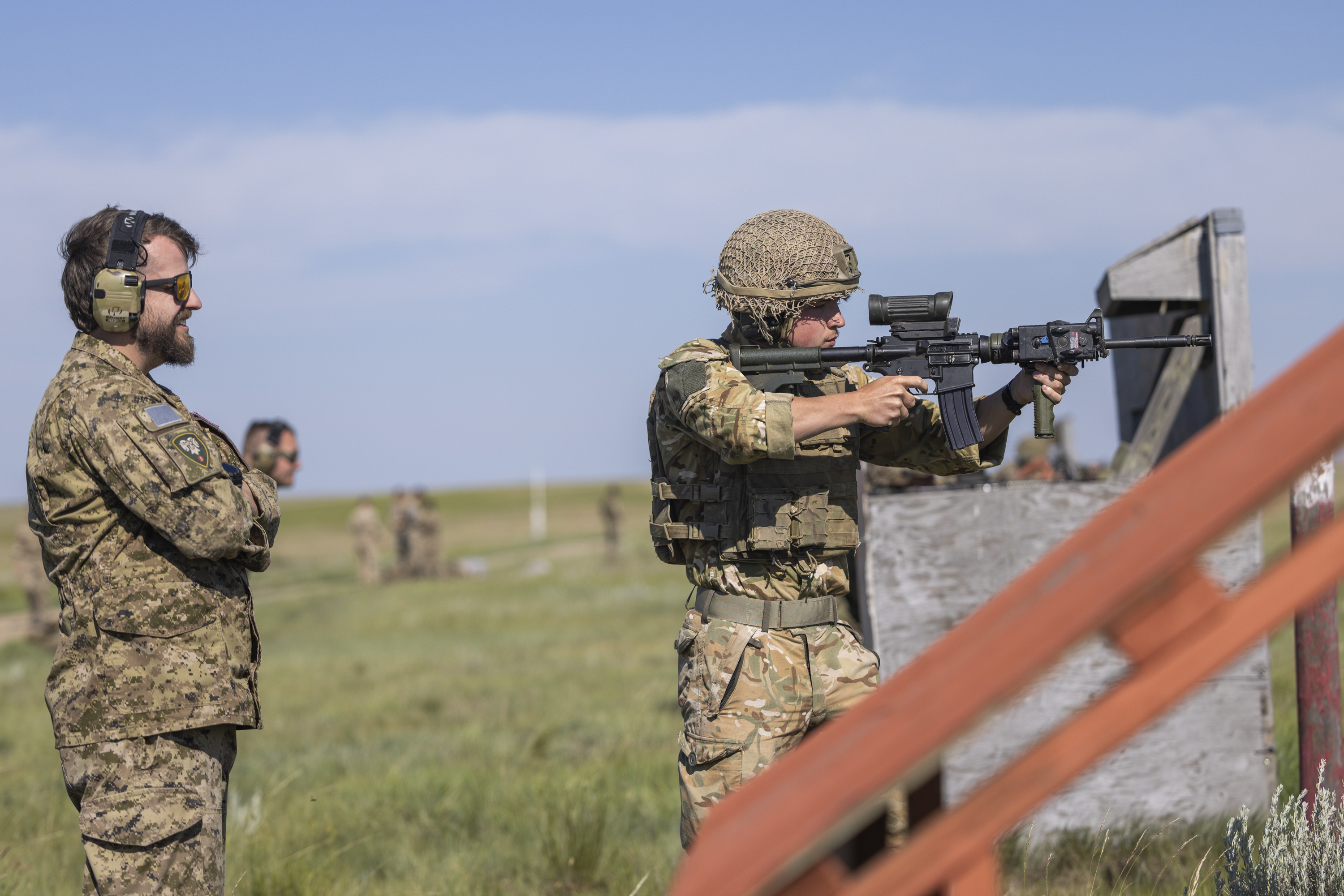 Two soldiers in camouflage uniforms are outdoors on a grassy field. One soldier is aiming a rifle while the other stands nearby observing. The scene is set under a clear blue sky.