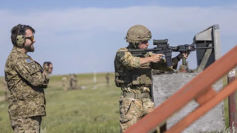 Two soldiers in camouflage uniforms are outdoors on a grassy field. One soldier is aiming a rifle while the other stands nearby observing. The scene is set under a clear blue sky.