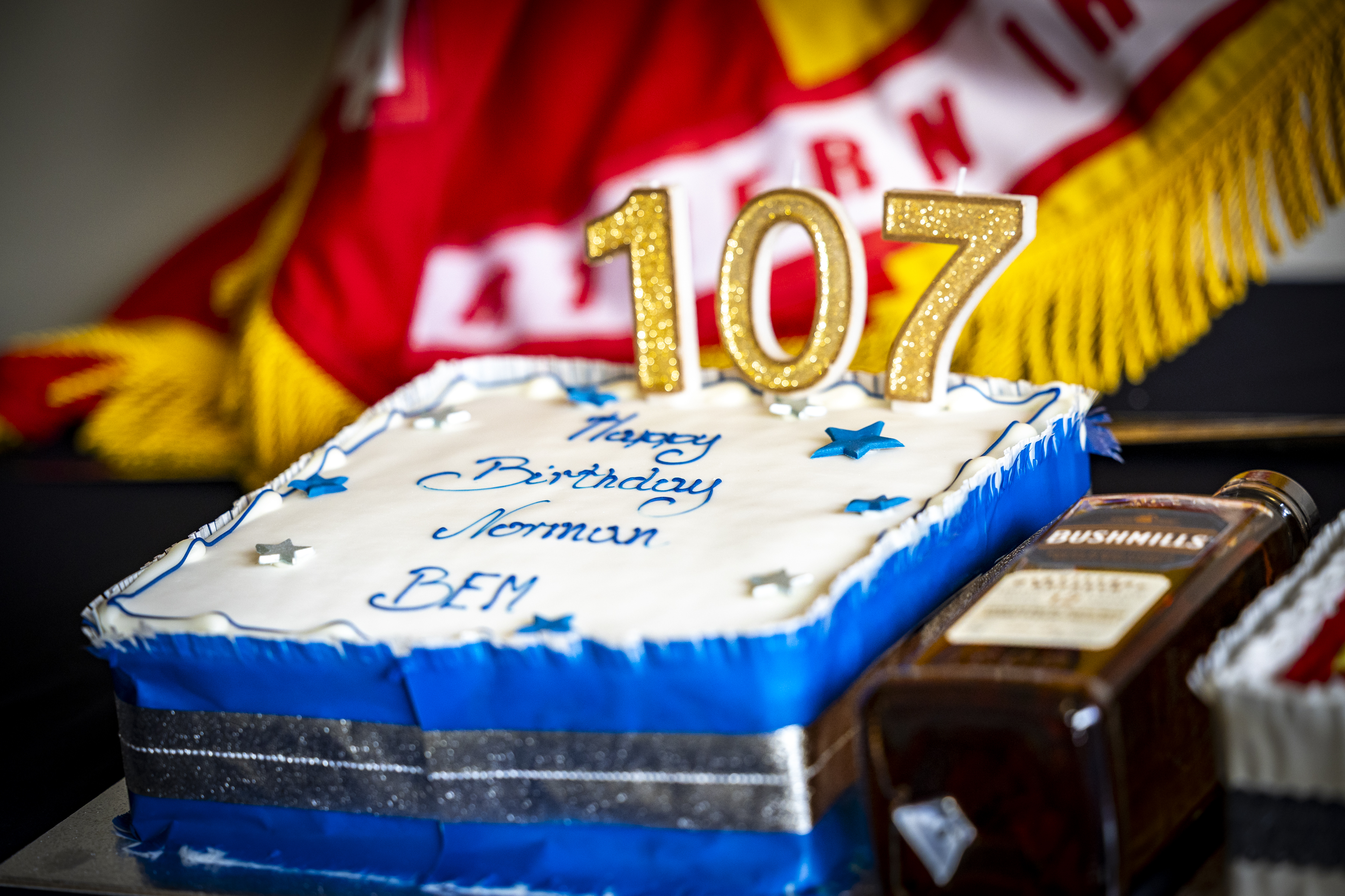 Rectangular birthday cake with blue and white icing, decorated with silver stars and gold candles forming the number 107, celebrating Norman's birthday.