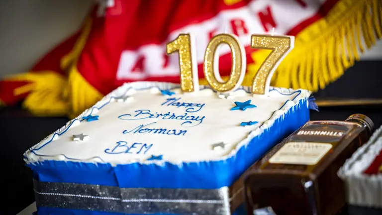 Rectangular birthday cake with blue and white icing, decorated with silver stars and gold candles forming the number 107, celebrating Norman's birthday.