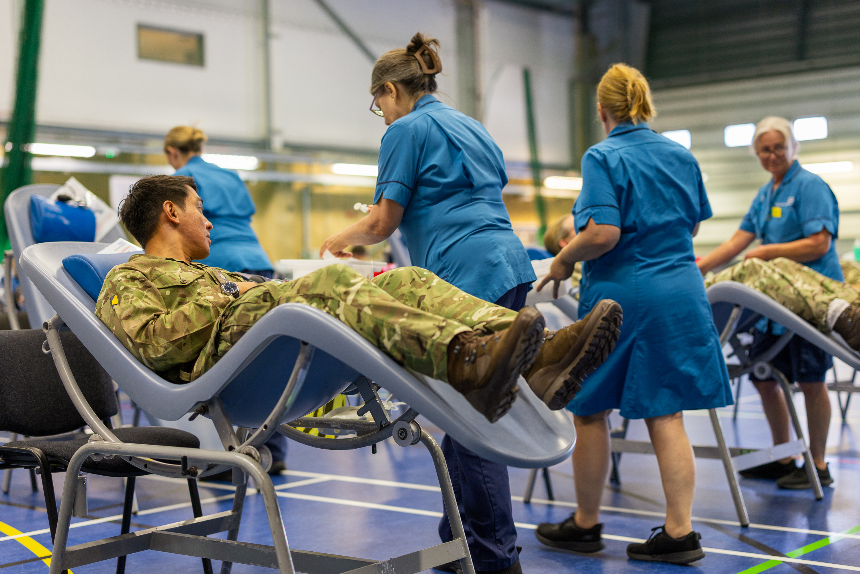 Military personnel lying on reclining chairs donating blood in a large indoor facility with nurses attending to them.
