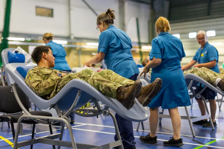 Military personnel lying on reclining chairs donating blood in a large indoor facility with nurses attending to them.