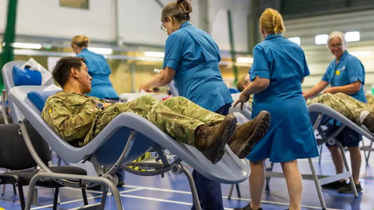 Military personnel lying on reclining chairs donating blood in a large indoor facility with nurses attending to them.