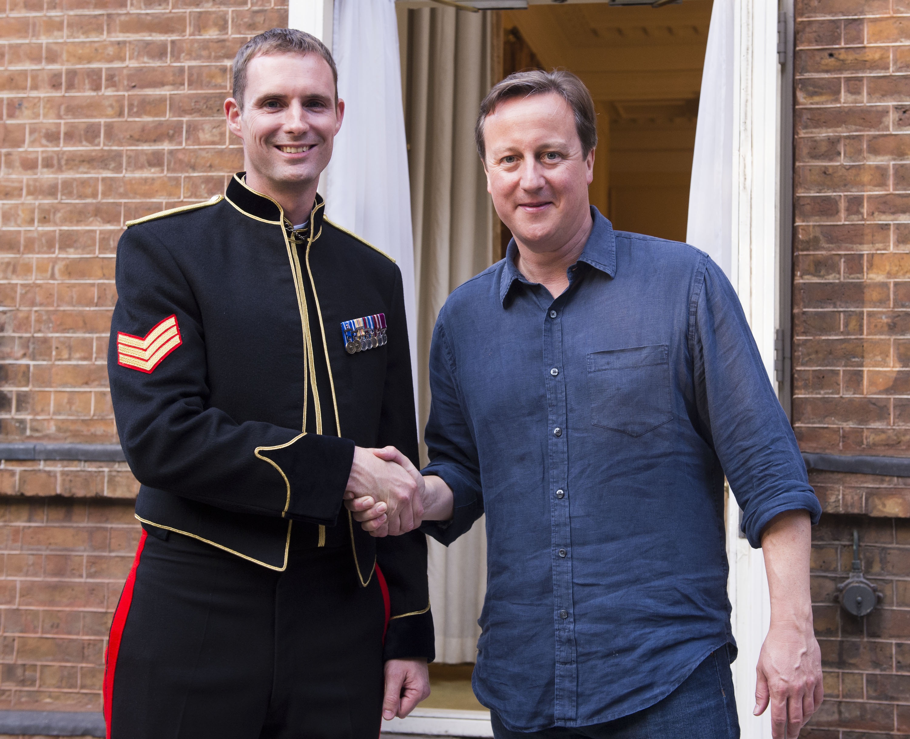A soldier in black ceremonial uniform stands shaking hands with former Prime Minister David Cameron.