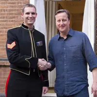 A soldier in black ceremonial uniform stands shaking hands with former Prime Minister David Cameron.