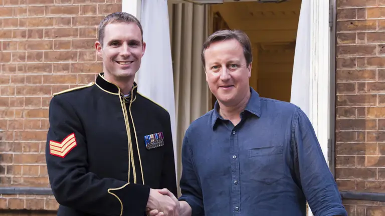 A soldier in black ceremonial uniform stands shaking hands with former Prime Minister David Cameron.