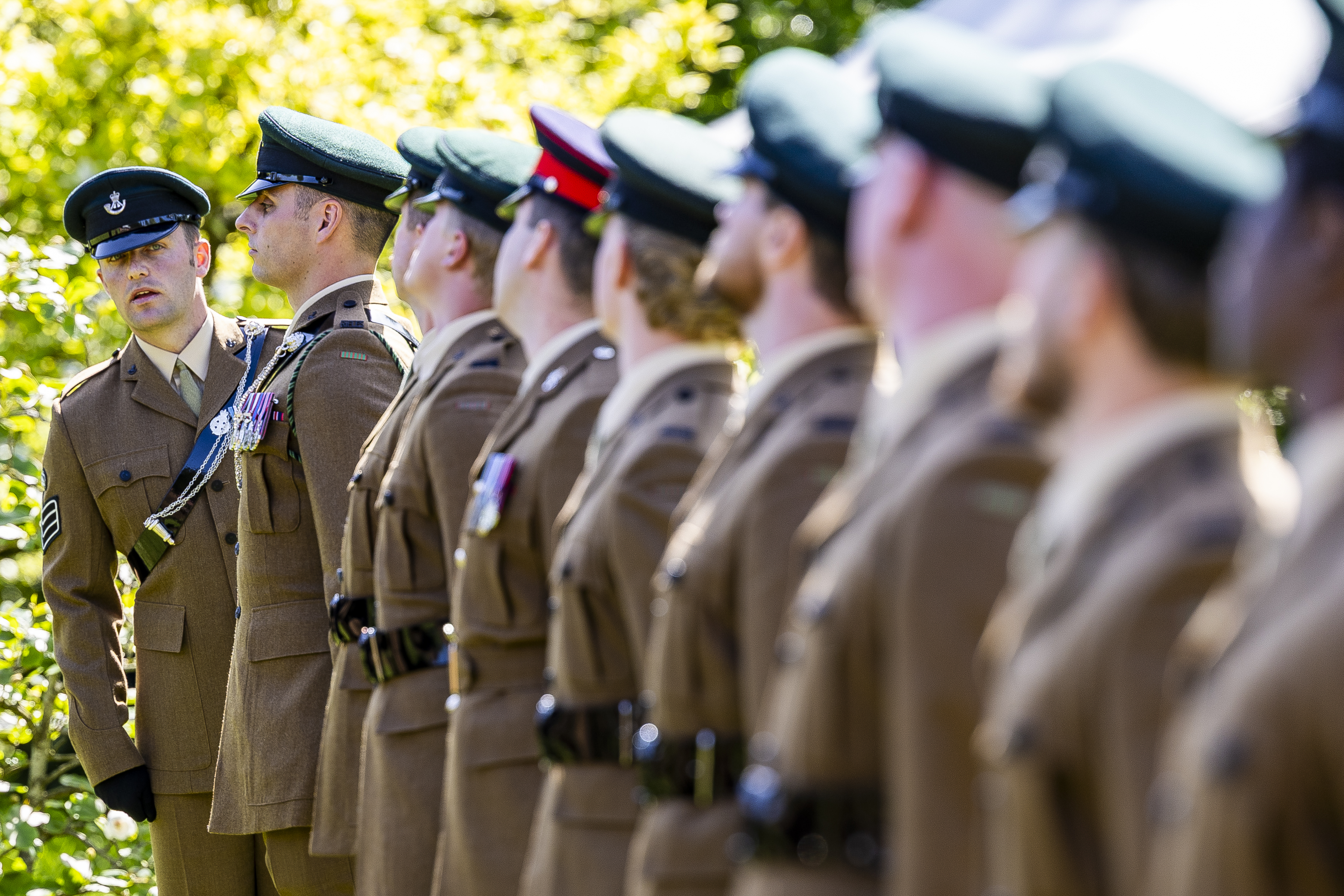A line of uniformed personnel stands at attention, wearing brown uniforms and green caps. The focus is on one person speaking to another in the row, surrounded by greenery. The setting appears formal and official.