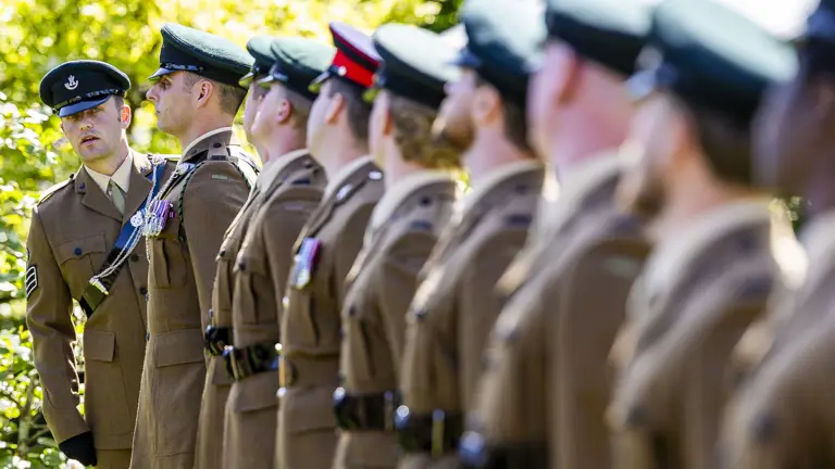 A line of uniformed personnel stands at attention, wearing brown uniforms and green caps. The focus is on one person speaking to another in the row, surrounded by greenery. The setting appears formal and official.