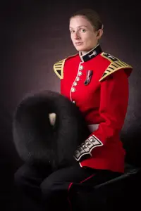 Female soldier is seen sat down posing for a professional portrait. She is wearing a red tunic and holding her black bear skin hat.