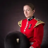 Female soldier is seen sat down posing for a professional portrait. She is wearing a red tunic and holding her black bear skin hat.