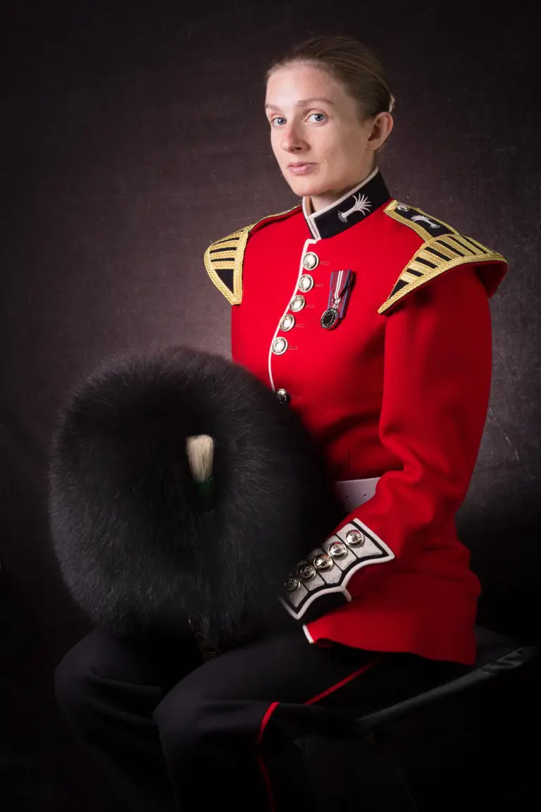Female soldier is seen sat down posing for a professional portrait. She is wearing a red tunic and holding her black bear skin hat.
