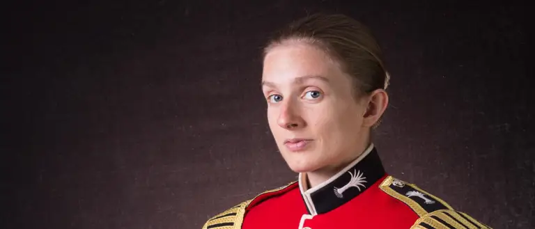 Female soldier is seen sat down posing for a professional portrait. She is wearing a red tunic and holding her black bear skin hat.