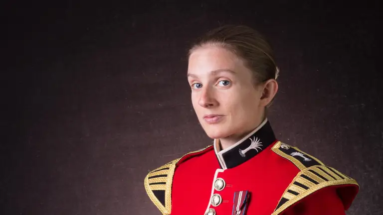 Female soldier is seen sat down posing for a professional portrait. She is wearing a red tunic and holding her black bear skin hat.