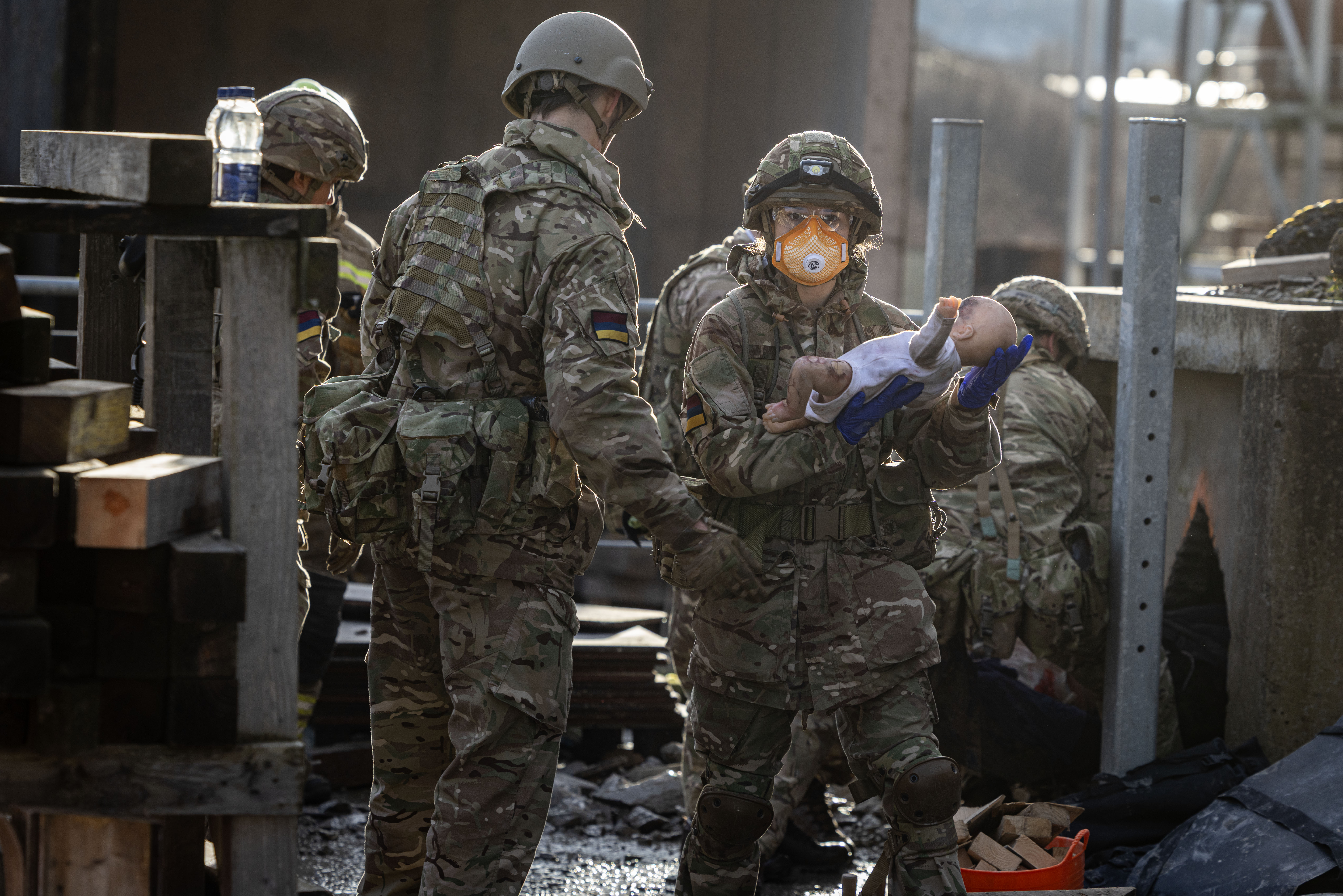 Soldiers in camouflage uniforms carry a doll through a damaged urban area during a rescue exercise.