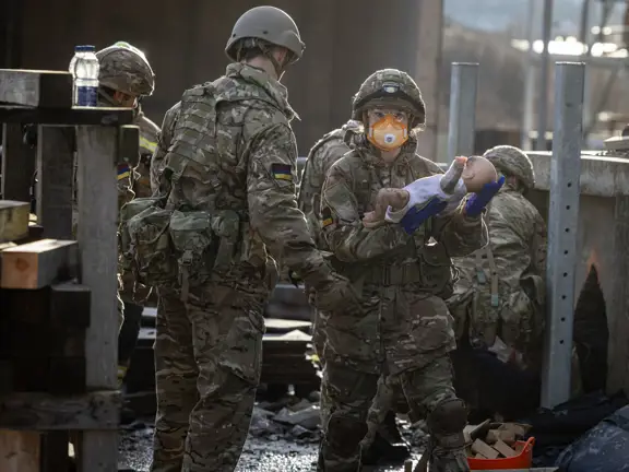 Soldiers in camouflage uniforms carry a doll through a damaged urban area during a rescue exercise.