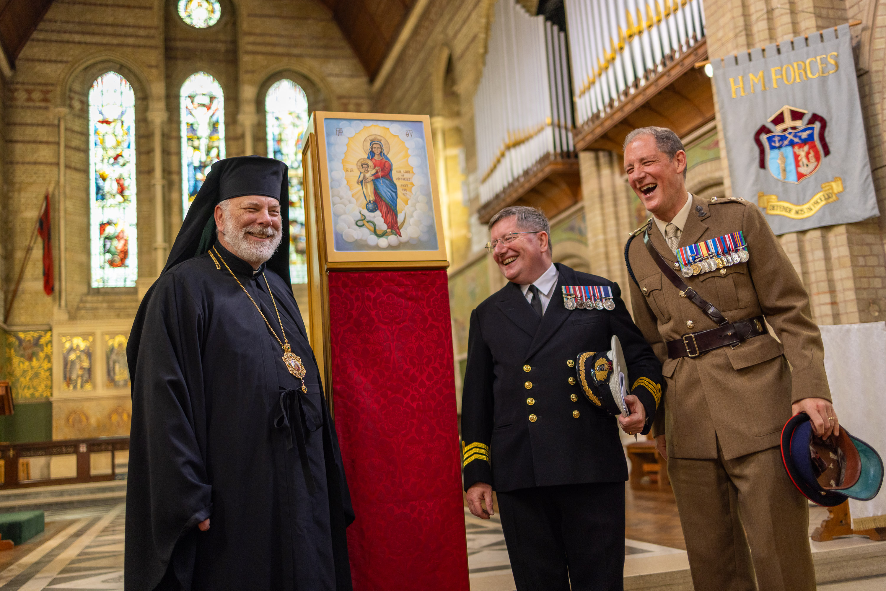 Three men in formal religious and military attire stand inside a church near a framed religious icon on a red pedestal.