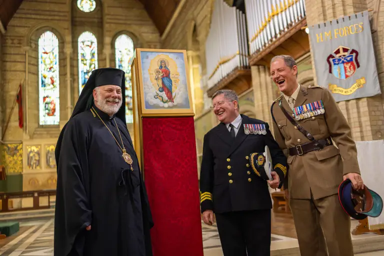 Three men in formal religious and military attire stand inside a church near a framed religious icon on a red pedestal.