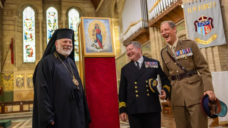 Three men in formal religious and military attire stand inside a church near a framed religious icon on a red pedestal.
