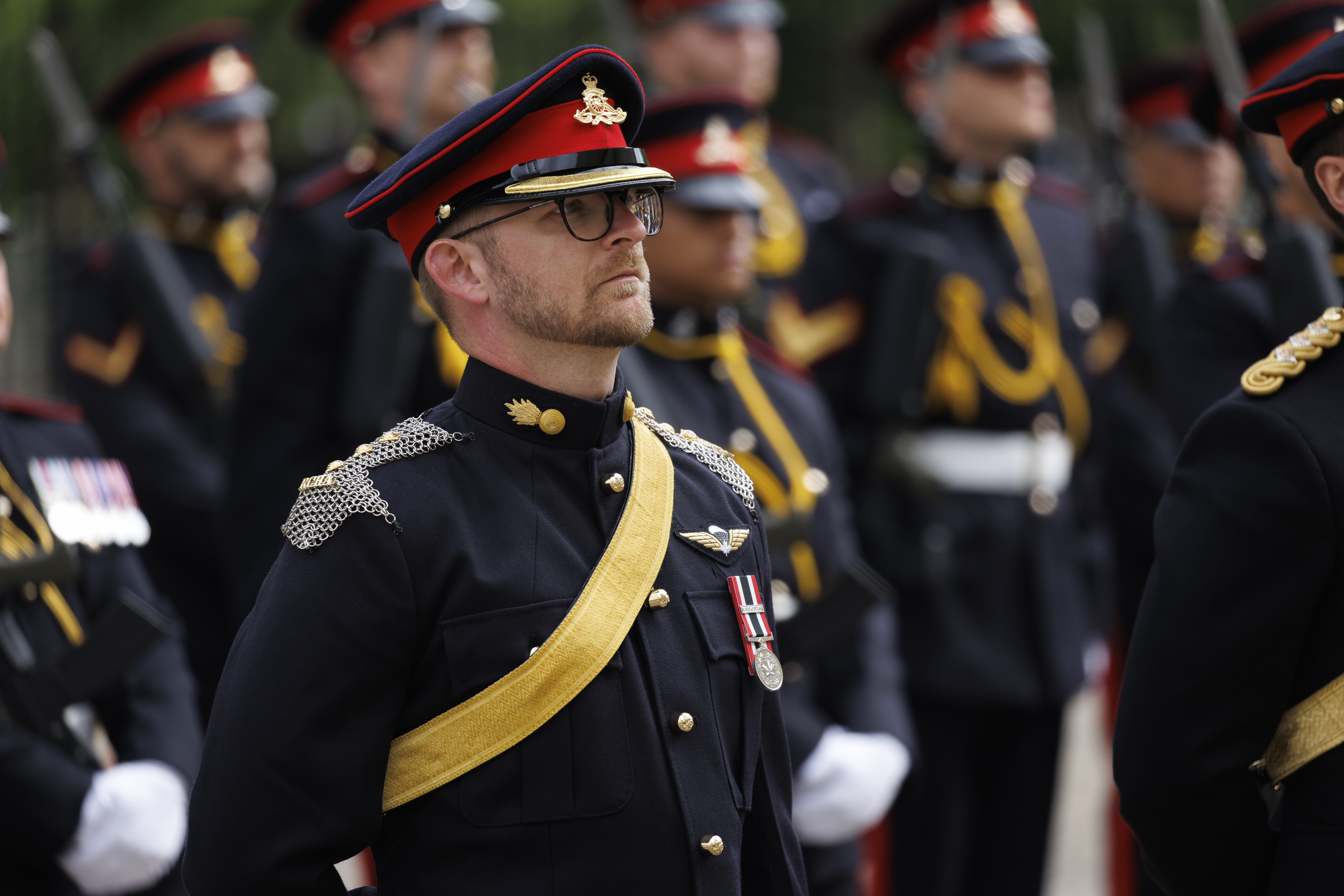 A group of individuals in formal military uniforms stands in formation. The uniforms are dark with red and gold accents.