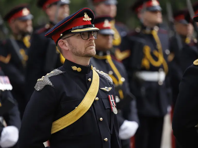 A group of individuals in formal military uniforms stands in formation. The uniforms are dark with red and gold accents.