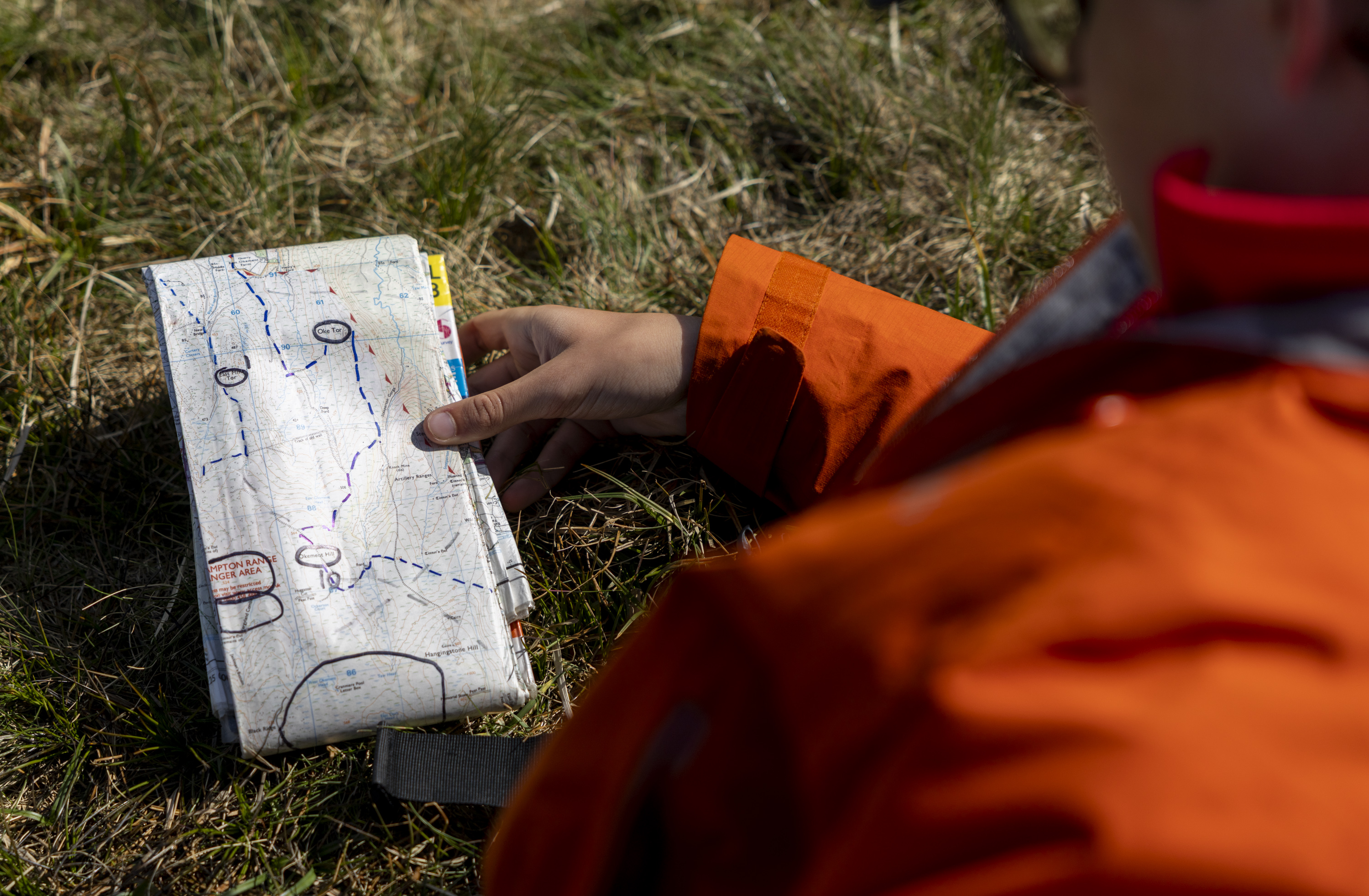 A person in an orange jacket sits on grass, holding an open map with marked routes. The scene suggests exploration and navigation.