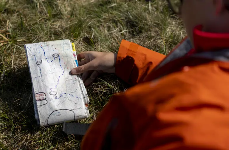 A person in an orange jacket sits on grass, holding an open map with marked routes. The scene suggests exploration and navigation.