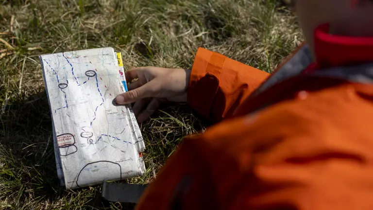 A person in an orange jacket sits on grass, holding an open map with marked routes. The scene suggests exploration and navigation.