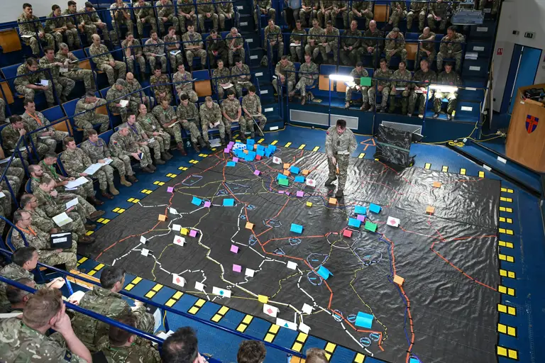 Soldiers sit around a ground map as a soldier stands on the map.