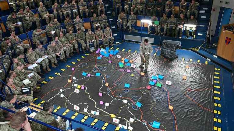 Soldiers sit around a ground map as a soldier stands on the map.