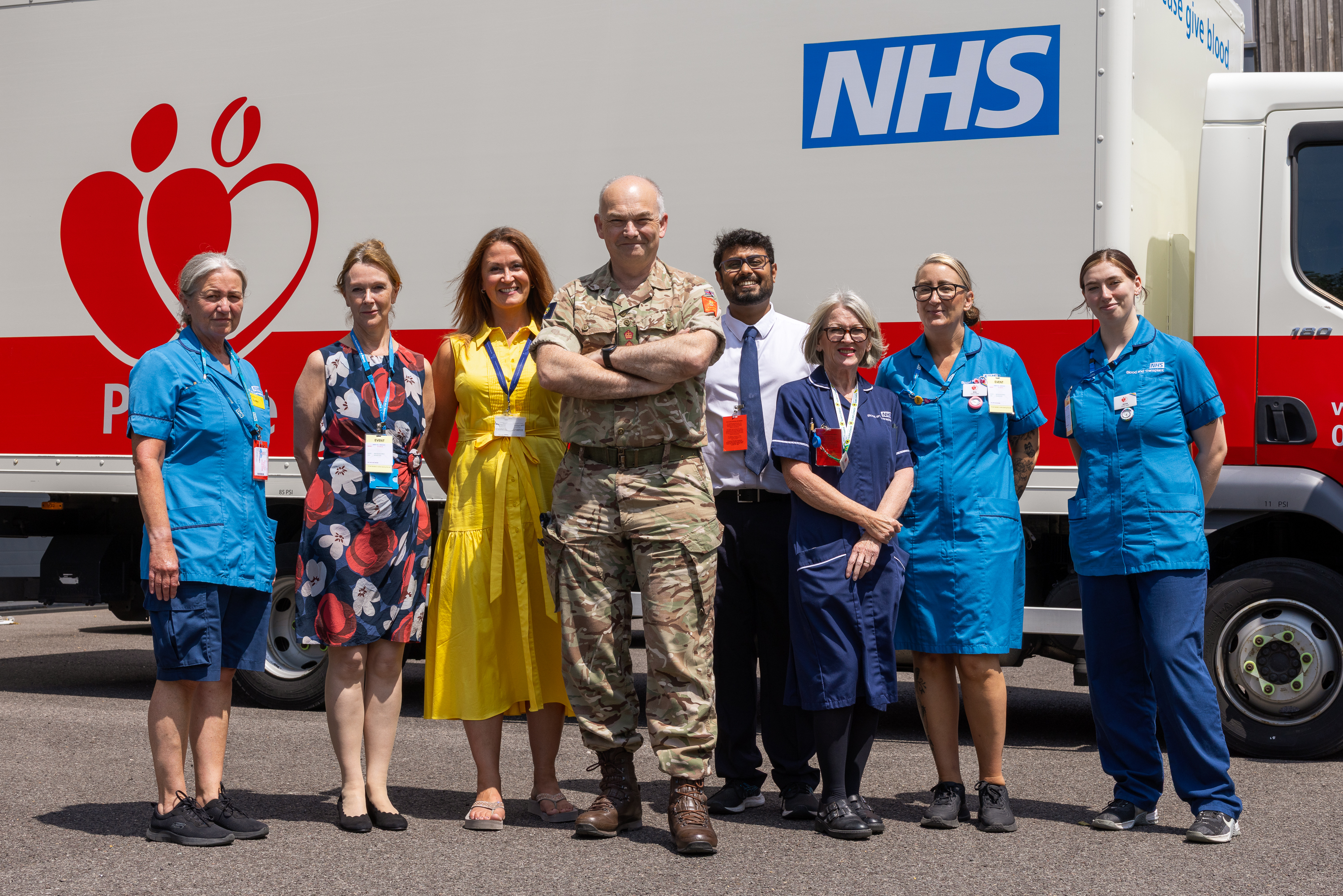 Group of NHS staff and a military personnel standing in front of a blood donation truck on a sunny day.